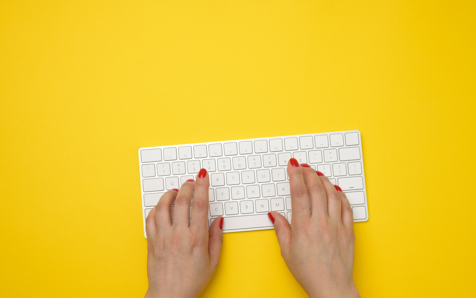 Two female hands press keys on a white wireless keyboard, yellow table